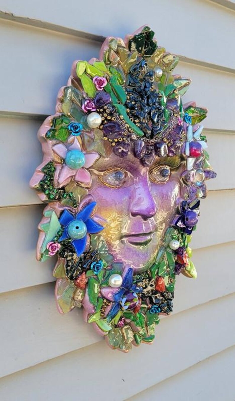 Puede incluir: Una m&aacute;scara decorativa de pared con un rostro central de color p&uacute;rpura iridiscente rodeado por un mosaico de fragmentos de vidrio verde, azul y rosa, peque&ntilde;as flores artificiales y cuentas de perlas blancas. La pieza tiene un aspecto texturizado.