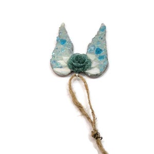 May include: A decorative ornament featuring a teal rose and two wing-shaped elements with a mosaic design in shades of blue and white. A natural twine loop is attached for hanging. The ornament is set against a white background.