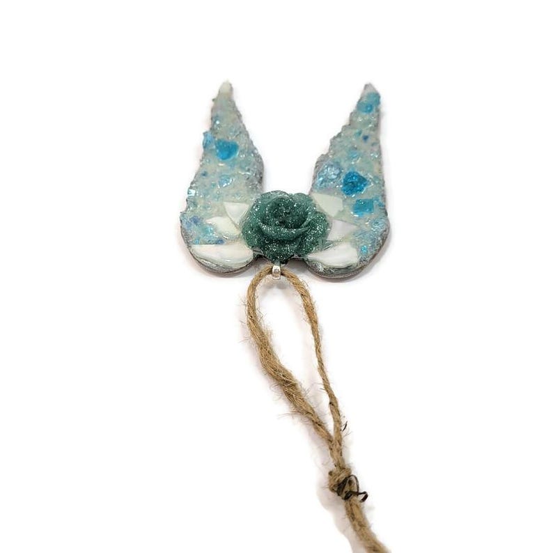 May include: A decorative ornament featuring a teal rose and two wing-shaped elements with a mosaic design in shades of blue and white. A natural twine loop is attached for hanging. The ornament is set against a white background.