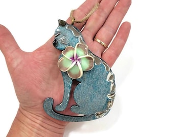 Op de afbeelding: Een blauw katvormig ornament met een glinsterende, glitterende afwerking en een lichtgroene en paarse plumeria-bloem op de borst. Het ornament is voorzien van een lus van jutekoord aan de bovenkant om op te hangen en heeft zilverkleurige accenten.