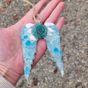 May include: A handcrafted angel wing ornament with a teal rose center. The wings are a mosaic of light blue and iridescent glass pieces, with a jute string for hanging. The ornament is designed for decorative purposes.