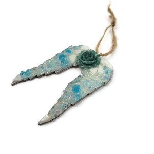 May include: A decorative angel wing ornament with a teal rose and a twine loop for hanging. The wings are light blue with iridescent glitter and teal accents. The ornament is on a white background.