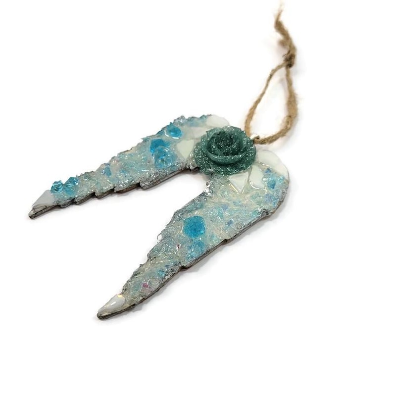 May include: A decorative angel wing ornament with a teal rose and a twine loop for hanging. The wings are light blue with iridescent glitter and teal accents. The ornament is on a white background.