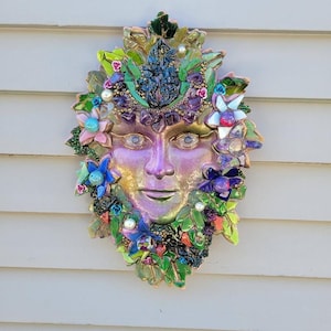 Puede incluir: Una escultura de m&aacute;scara para pared con un rostro central iridiscente rodeado por un mosaico de hojas verdes, piedras moradas, flores azules y rosas, y peque&ntilde;as perlas blancas. La pieza decorativa muestra una vibrante gama de colores.