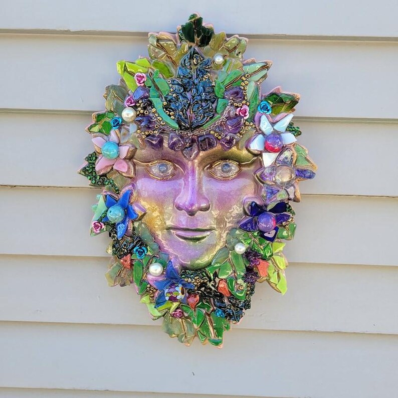 Puede incluir: Una escultura de m&aacute;scara para pared con un rostro central iridiscente rodeado por un mosaico de hojas verdes, piedras moradas, flores azules y rosas, y peque&ntilde;as perlas blancas. La pieza decorativa muestra una vibrante gama de colores.