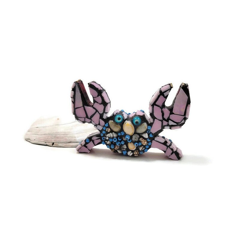 May include: A pink and blue mosaic crab sculpture with shell and bead accents. The crab is standing on a piece of driftwood.