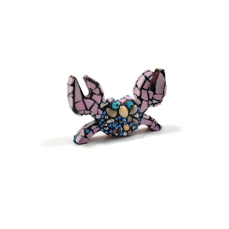 May include: A pink mosaic crab figurine with blue and white eyes. The crab is made of small pieces of glass and shell.