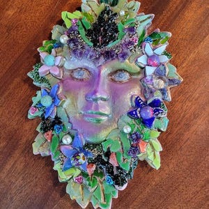 Puede incluir: Una m&aacute;scara decorativa de pared con un rostro central de acabado iridiscente en tonos morados y verdes. La m&aacute;scara est&aacute; rodeada por un mosaico de hojas de vidrio verde, piedras de amatista moradas, peque&ntilde;as perlas blancas y flores variadas.