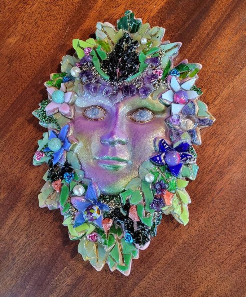 Puede incluir: Una m&aacute;scara decorativa de pared con un rostro central de acabado iridiscente en tonos morados y verdes. La m&aacute;scara est&aacute; rodeada por un mosaico de hojas de vidrio verde, piedras de amatista moradas, peque&ntilde;as perlas blancas y flores variadas.