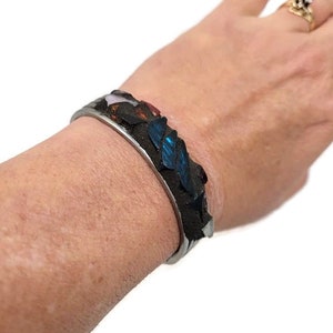 May include: A silver bracelet with a mosaic design featuring blue, red, and white glass pieces. The bracelet is made of metal and has a smooth, polished finish.