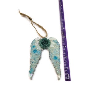May include: A decorative angel wing ornament with a teal rose in the center. The wings are light blue and white with iridescent accents. The ornament is approximately 4 inches long and hangs from a natural twine loop.