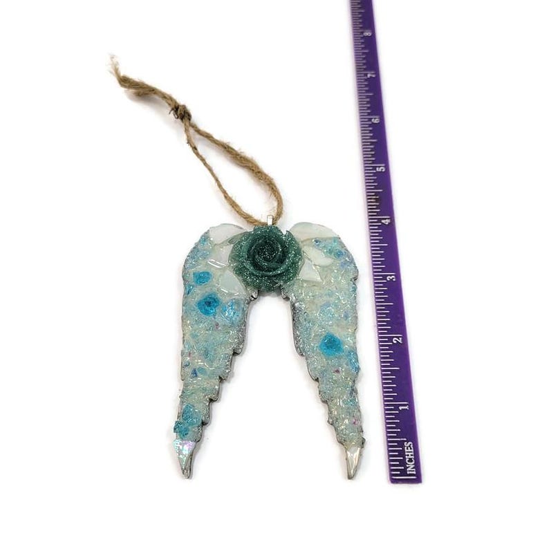 May include: A decorative angel wing ornament with a teal rose in the center. The wings are light blue and white with iridescent accents. The ornament is approximately 4 inches long and hangs from a natural twine loop.
