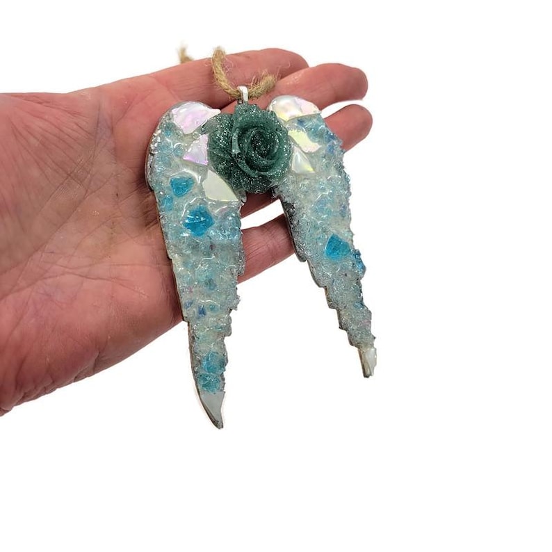 May include: A decorative angel wing ornament with a teal rose center. The wings are made of mosaic glass in shades of blue and white, with iridescent accents. A jute string is attached for hanging.