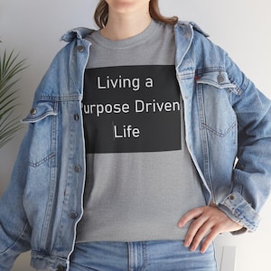 以下が含まれることがあります： 胸元に黒い長方形のグラフィックと白い文字でLiving a Purpose Driven LifeとプリントされたヘザーグレーのTシャツ。金属製のボタンが付いたライトブルーのデニムジャケットとブルーのデニムパンツを着用。