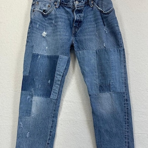 Puede incluir: Pantalones vaqueros de mezclilla azul con un diseño de parches en diferentes tonos de azul. Los pantalones incluyen cierre de botones, trabillas para cinturón, cinco bolsillos y zonas desgastadas con pequeñas secciones deshilachadas.