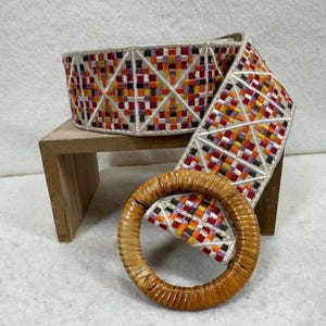 May include: A woven belt featuring a geometric pattern of orange, red, purple, and white threads. The belt is finished with a large, circular, light brown rattan buckle. The design consists of small, repeating squares arranged in a grid.