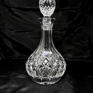 May include: A clear crystal decanter featuring a bulbous base with a diamond-cut pattern and a matching faceted stopper. A small blue and gold circular sticker with the text Bohemia is affixed to the neck of the decanter, which has a smooth, tapered neck leading to a flared rim.