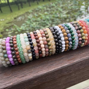 Può includere: Una collezione di braccialetti elastici con perline di pietre preziose rotonde e levigate in un'ampia varietà di colori, tra cui viola, verde, nero, bianco, rosa, blu e arancione, esposti su un supporto cilindrico bianco e dorato.