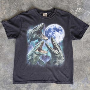 May include: A black short-sleeve t-shirt featuring a graphic print of three mosasaurs with open mouths positioned beneath a large, textured full moon. The design includes blue and purple nebula-like accents surrounding the prehistoric marine reptiles.