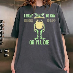 Puede incluir: Una camiseta de manga corta gris carbón con un gráfico de una rana verde con vientre amarillo. El texto impreso en verde y beige dice: I HAVE WEIRD STUFF TO SAY WEIRD STUFF OR I'LL DIE. La camiseta tiene cuello redondo y un corte holgado.