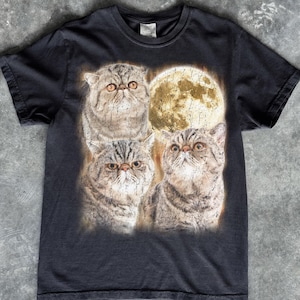 May include: A black short-sleeve t-shirt featuring a graphic print of three brown tabby cats with flat faces arranged around a large, textured, glowing yellow full moon. The cats have wide, alert eyes and detailed fur patterns.