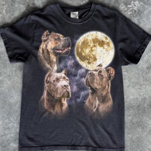 May include: A black short-sleeve t-shirt featuring a graphic print of three brown Cane Corso dogs arranged around a large, textured full moon. The background includes dark, cloudy, and purple-toned atmospheric elements surrounding the dogs.