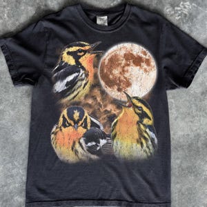 May include: A black short-sleeved t-shirt featuring a graphic print of three yellow and black birds positioned around a large, textured, circular moon. The birds are depicted in various poses against a dark, cloudy, and atmospheric background.