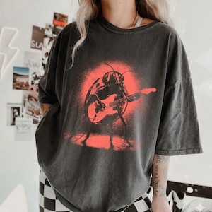 Pode incluir: Uma t-shirt cinzento-carvão de corte oversize com uma estampa gráfica de um escaravelho a tocar uma guitarra elétrica. O escaravelho está centrado num efeito de tinta spray circular vermelho vivo. A t-shirt tem mangas curtas e gola redonda.