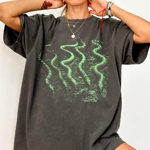 May include: A charcoal grey short-sleeve t-shirt featuring a graphic print of four wavy, bright green snakes positioned vertically across the centre. The shirt has a relaxed, loose fit with a crew neckline and a faded, vintage-style fabric finish.