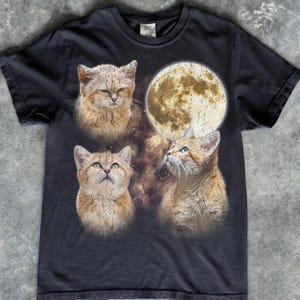 May include: A black short-sleeved t-shirt featuring a graphic print of three tan cats and a large, textured full moon. The cats are positioned around the moon against a dark, nebulous background, with a distressed, vintage-style crackle effect.