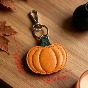 May include: A pumpkin-shaped keychain with orange textured material and a dark green stem. It features a silver metal lobster clasp and a split ring. Red text indicates dimensions of 3.15 inches in height and 2.56 inches in width.