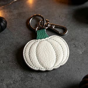 May include: A white pumpkin-shaped keychain made of textured leather with a green stem attachment. The keychain features a silver-toned metal split ring and a lobster clasp for secure attachment to keys or bags, showcasing detailed stitching.