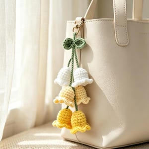 May include: A cream-colored leather handbag featuring a crochet flower keychain. The keychain consists of two green stems with leaves at the top, each ending in three stacked bell-shaped flowers in white, pale yellow, and bright yellow.