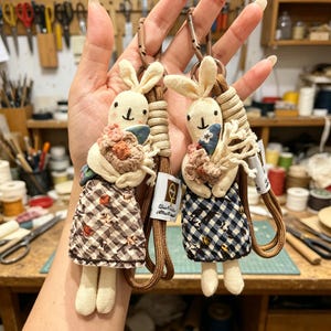 May include: Two fabric rabbit keychains with embroidered faces, each holding a small crocheted flower bouquet. One rabbit wears a brown and white checkered dress, the other a navy and white checkered dress. Both feature a brown cord and metal clip.