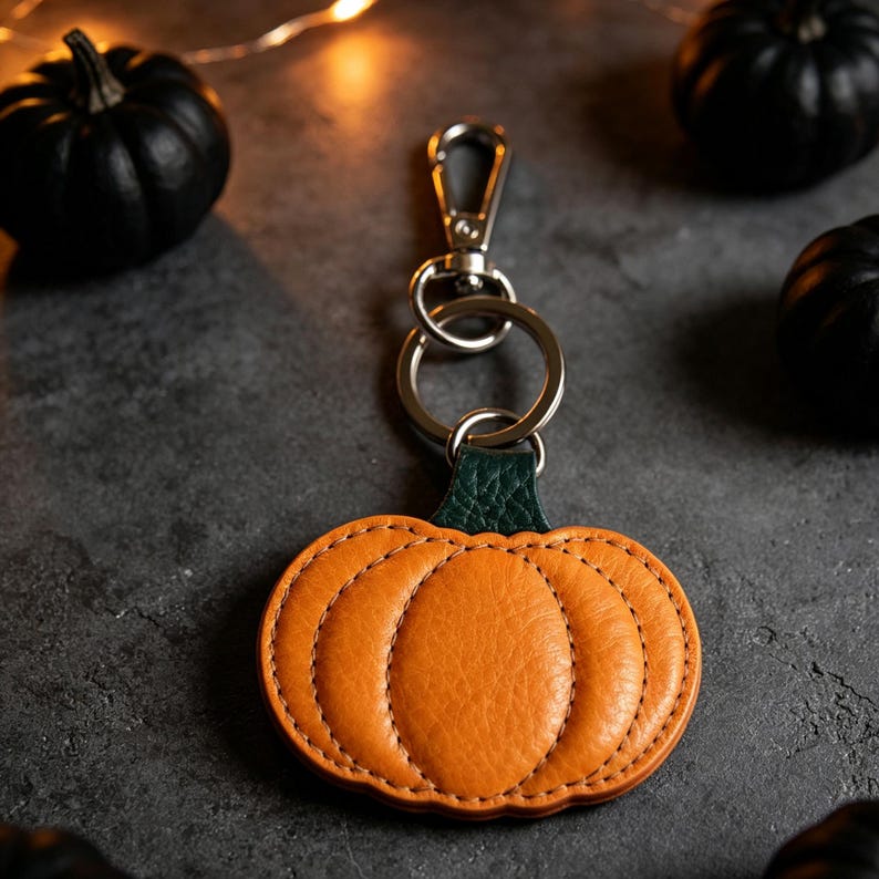 May include: A pumpkin-shaped keychain crafted from orange textured material with a dark green stem. The pumpkin is attached to a silver-toned metal split ring and a lobster clasp. The design features visible stitching along the pumpkin segments.
