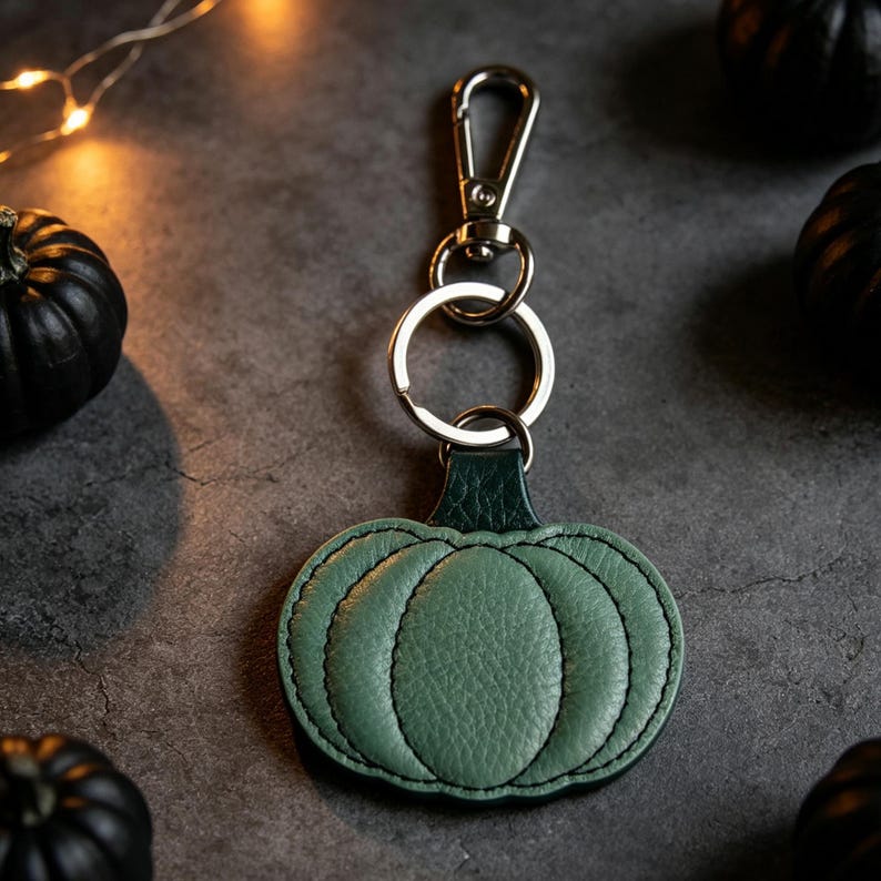 May include: A sage green leather pumpkin keychain featuring a dark green stem, stitched detailing to define the pumpkin segments, and a silver-toned metal split ring attached to a lobster claw clasp for secure fastening to keys or bags.