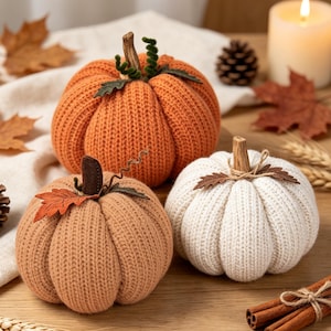May include: Three decorative pumpkins crafted from knit fabric in orange, beige, and white. Each pumpkin features a textured ribbed surface, a brown stem, and small fabric leaf accents. The orange pumpkin includes green curly tendrils.