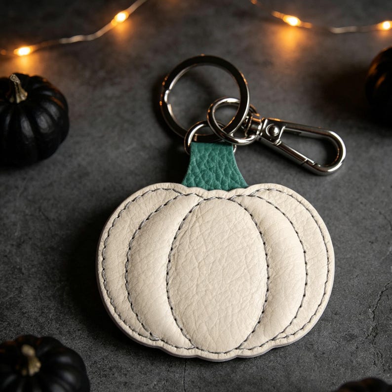 May include: A pumpkin-shaped keychain crafted from off-white textured leather with a green leather stem. The pumpkin features visible stitching along its segments and is attached to a silver-toned metal split ring and a lobster claw clasp.