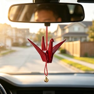 May include: A red leather origami crane hanging ornament suspended from a thin red cord. The crane features stitched detailing and is attached to a small, round, metallic gold-coloured bell at the bottom, designed for display in a vehicle.