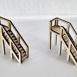 May include: Two miniature staircases constructed from light-colored wood with dark, laser-cut edges. Each structure features a raised platform, a set of steps leading down, and side railings with vertical supports, creating a detailed architectural model.