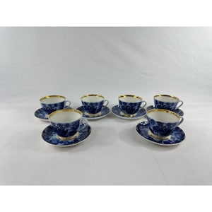 May include: A set of six porcelain teacups and matching saucers featuring a deep blue floral pattern on a white background. Each cup and saucer is finished with a thin gold rim along the edges, providing a consistent look across the entire collection.