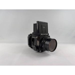 May include: A black Mamiya RB67 medium format film camera featuring a modular body design, a waist-level viewfinder, and a lens with a prominent front element. The camera body displays the Mamiya RB67 logo on the front panel and includes various manual adjustment dials and controls on the side.