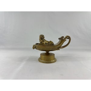 May include: A metallic gold-toned oil lamp featuring a sculpted lion figure resting on the top surface. The lamp has a curved handle, a fluted bowl design, and sits on a circular pedestal base with decorative detailing around the bottom edge.