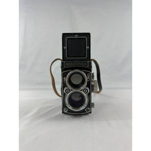 May include: A black Rolleiflex twin-lens reflex camera featuring a top-mounted viewfinder hood, two circular lenses stacked vertically, and a brown leather neck strap attached to the sides. The camera body displays the brand name Rolleiflex.