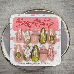 May include: A set of ten almond-shaped press-on nails on a Cherry Nail Co display card. The nails feature a light pink base with decorative elements including white floral designs, metallic rose gold accents, green glitter, and small pearls.