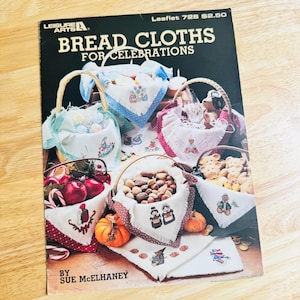 May include: A Leisure Arts craft booklet titled Bread Cloths for Celebrations by Sue McElhaney. The cover features multiple woven baskets lined with white fabric cloths, each decorated with various embroidered designs and fringed edges.