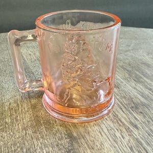 May include: A translucent pink glass mug featuring a molded relief of a figure playing a pipe. The text TOM TOM THE PIPER'S SON is embossed on the front surface. The mug has a sturdy handle and a smooth, circular base, showcasing a vintage design.