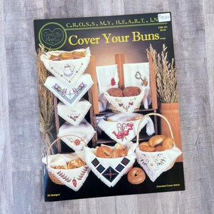 May include: A Cross My Heart Inc. pattern book titled Cover Your Buns featuring 22 counted cross stitch designs. The cover displays various white fabric cloths with embroidered motifs used to line baskets and cover baked goods like buns.
