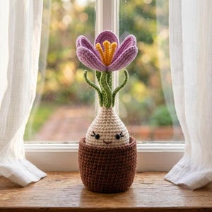 以下が含まれることがあります： 紫色の花びらと黄色の雄しべを持つかぎ針編みのクロッカスの花。クリーム色の球根の土台には、刺繍された笑顔が施されています。球根は質感のある茶色のかぎ針編みの鉢に入っており、手作りの装飾品となっています。