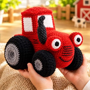 Puede incluir: Un juguete de tractor tejido a ganchillo en color rojo con ventanas blancas, un tubo de escape negro y grandes ruedas negras con centros grises. El tractor tiene dos ojos redondos blancos con pupilas negras y una sonrisa negra bordada.